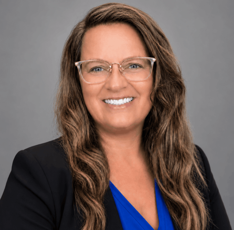 Ashley Jean Gorby wearing a blue shirt with black jacket and glasses with gray background